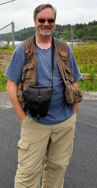 Jay Brass standing at St. Johns Bay, wearing a photographer's vest with a camera bag slung across his chest.