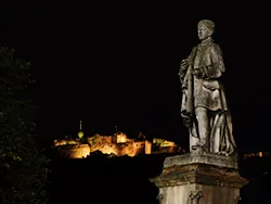 Statue lit from the front at night, gazing into the frame, with Edinburgh Castle behind it
