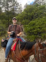 Jay Brass on horseback, camera in hand, riding a trail through the pines in Colorado.