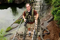 Jay Brass crossing a rope-and-plank footbridge over a river, carrying his camera gear, with alligators resting below.