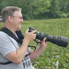 Jay Brass holding a camera fitted with a long telephoto lens in a green field upstate.